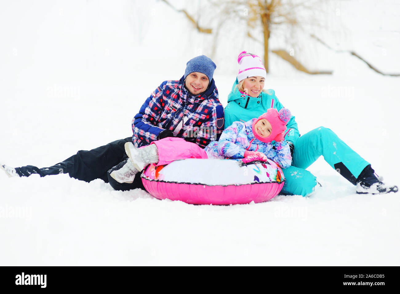 Eine junge Familie - Mutter, Vater und Tochter in hellen Skianzuege sitzen auf dem Schnee mit Schlauch oder aufblasbare Schlitten. Winter Unterhaltung Stockfoto
