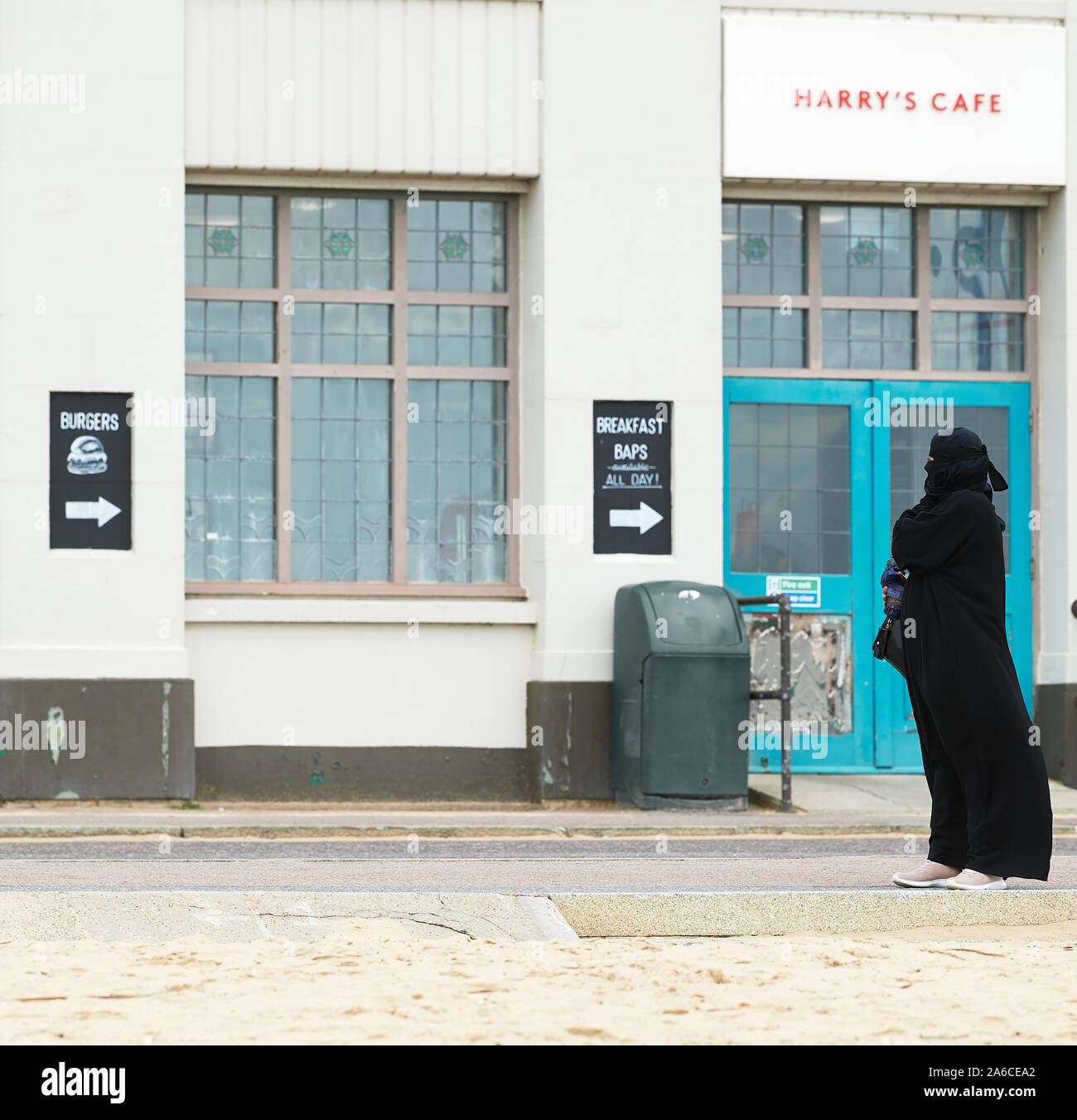 Eine Frau in voller nikab Hijab schwarzen Kleid steht auf der Promenade nahe der Sandstrand in Bournemouth, England, außerhalb von Harry's Cafe. Stockfoto