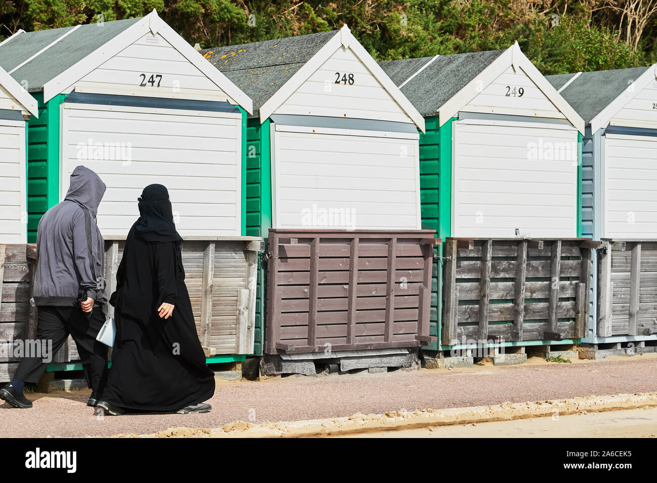 Eine Frau in voller Schwarz nikab hijab Spaziergänge entlang der Promenade mit ihrem Ehemann Vergangenheit Strand Hütten neben der Sandstrand in Bournemouth, England. Stockfoto