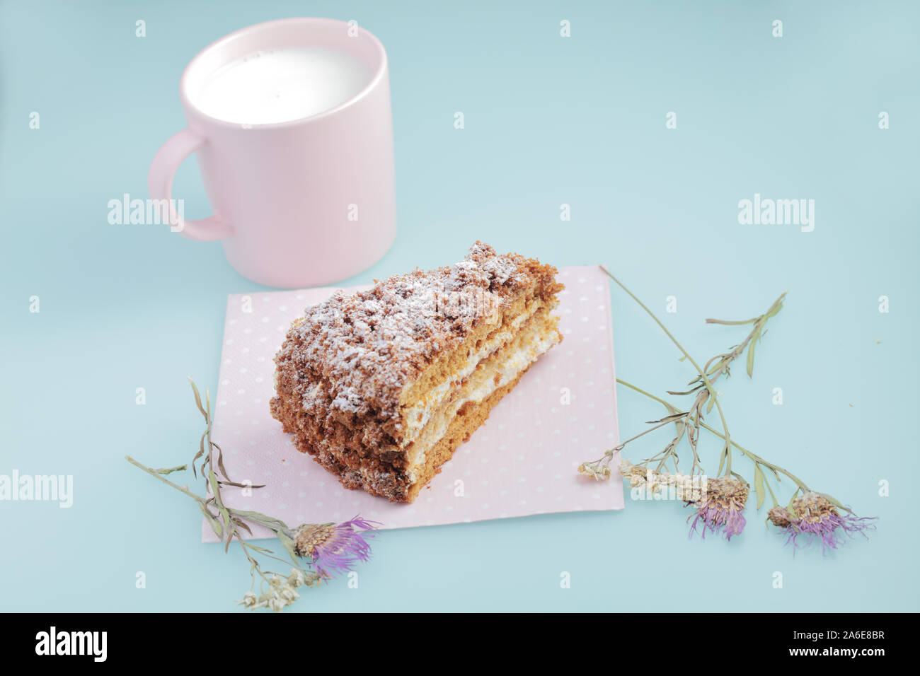 Scheibe russischer Kuchen medovik mit Glasur auf Enteneier blauem Hintergrund mit getrockneten Blumen und rosa Tasse Milch. Pastellfarben und Copyspace. Stockfoto