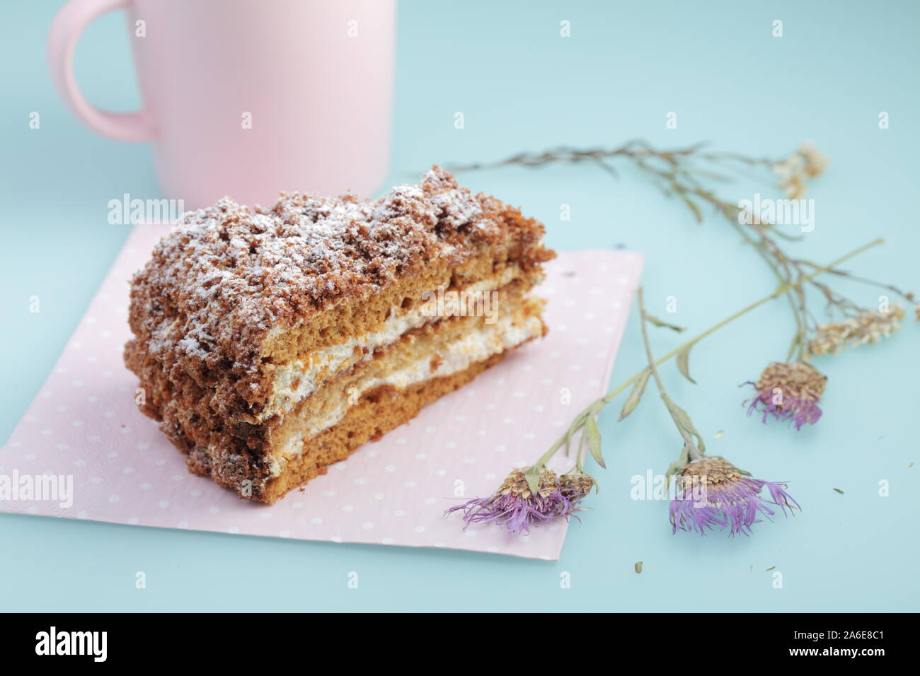 Scheibe russischer Kuchen medovik mit Glasur auf Enteneier blauem Hintergrund mit getrockneten Blumen und rosa Tasse Milch. Pastellfarben und Copyspace. Stockfoto