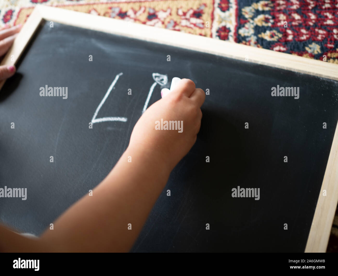 4-Jähriges Mädchen schreibt Zeichen L und I auf einer Schiefertafel, Teppich im Hintergrund. Stockfoto