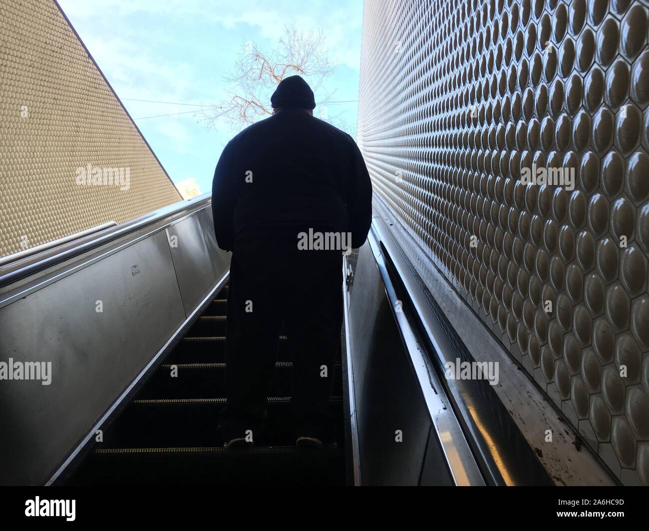 Silhouette eines großen Mannes mit abgerundeten Schultern stehend auf einer Rolltreppe mit strukturierter Wand, Treppen gehen sie nach oben in Richtung der Freien und blauer Himmel Stockfoto