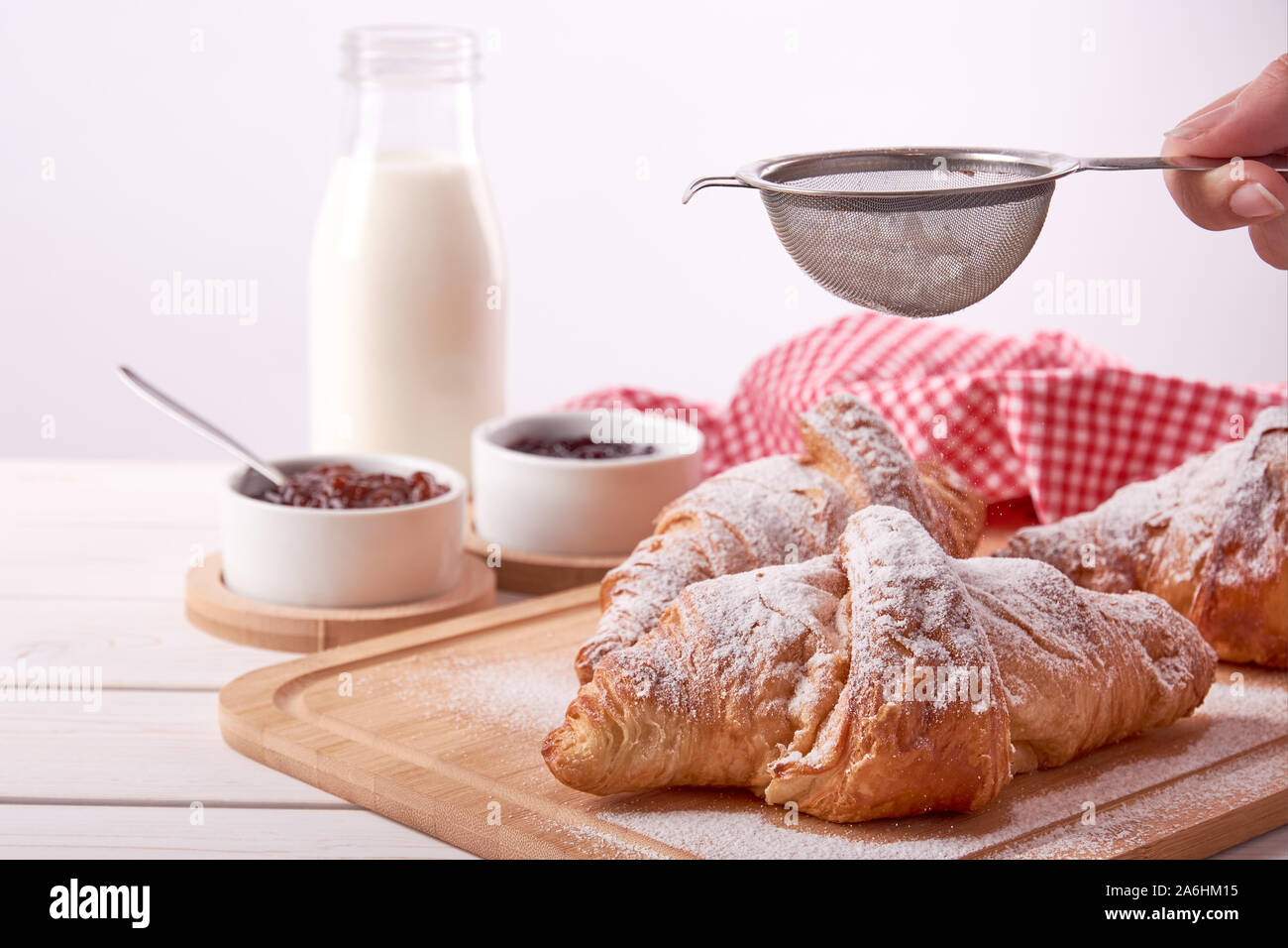 Stillleben mit gezuckertem Croissant, Porzellan Schüsseln mit Marmelade und ein Glas Milch und weibliche Hand mit Zucker, auf weißem Holz- Tisch mit Platz f Stockfoto