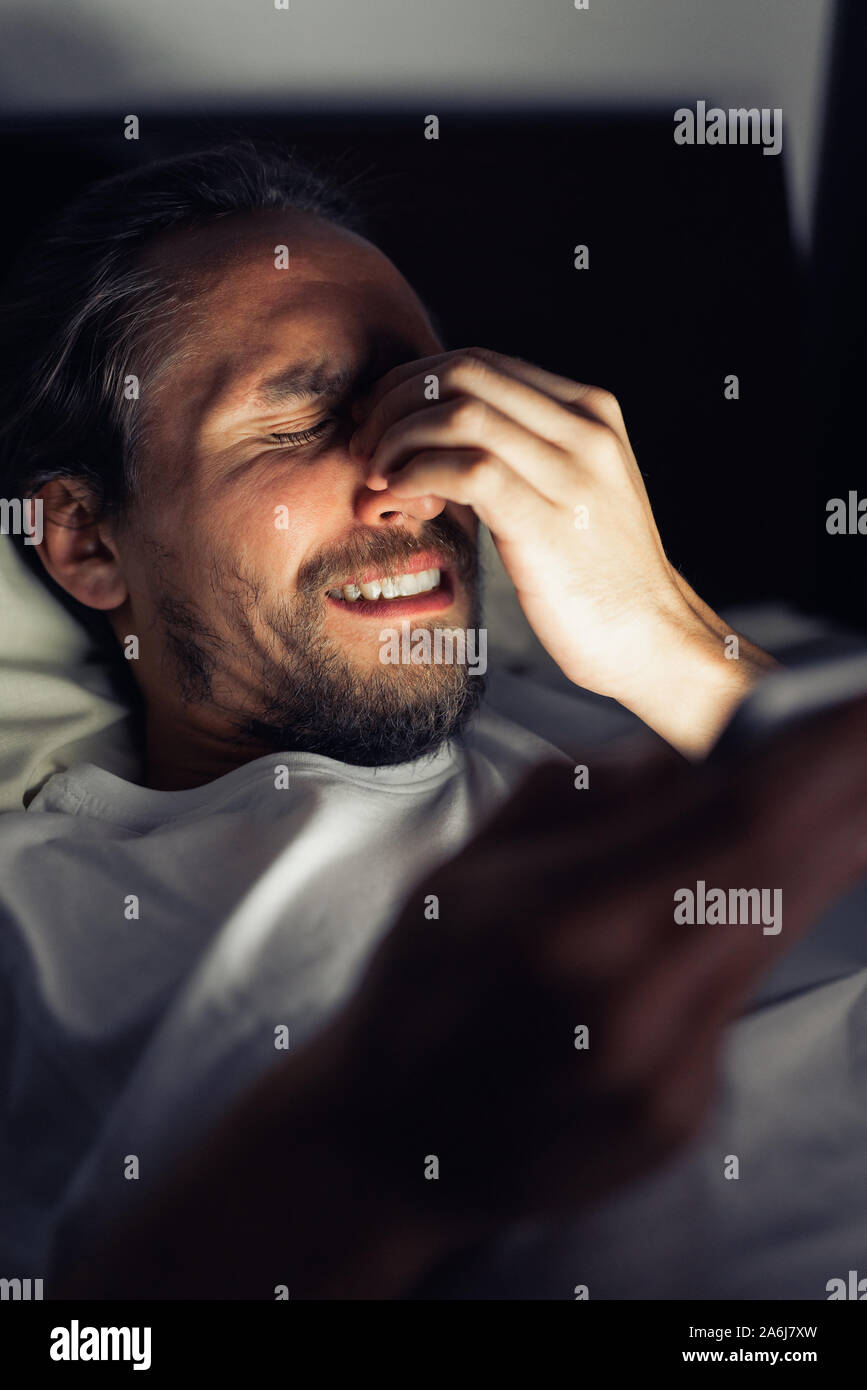 Bärtige Kaukasier Männlich ist etwas aufpassen auf seinem Tablet nachts im Bett. Er ist müde, kann nicht schlafen und er hat Kopfschmerzen. Stockfoto