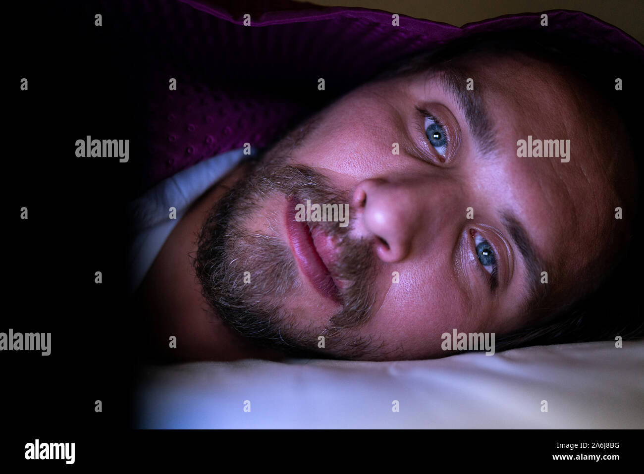 Bärtiger junger Mann im Bett liegend unter seiner Decke. Er kann nicht schlafen und ist etwas aufpassen auf sein Mobiltelefon. Stockfoto