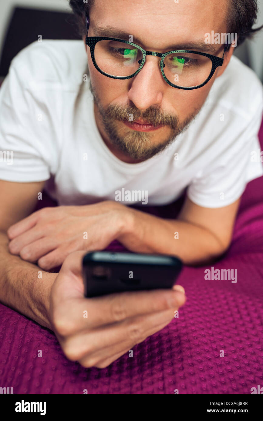 Foto von einem eher ernsten jungen gutaussehenden Mann der Interaktion mit seinem mobilen Gerät, während Sie im Bett lag, Stockfoto