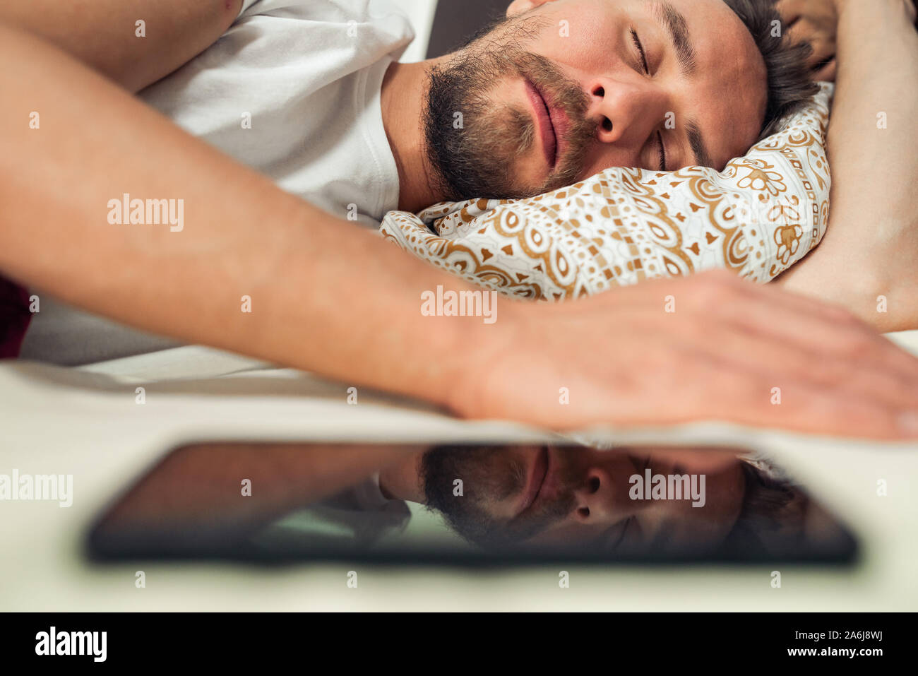 Telefon im Bett neben einem gutaussehenden jungen Mann, dass ist ein Nickerchen. Reflexion des Telefons zu sehen. Stockfoto
