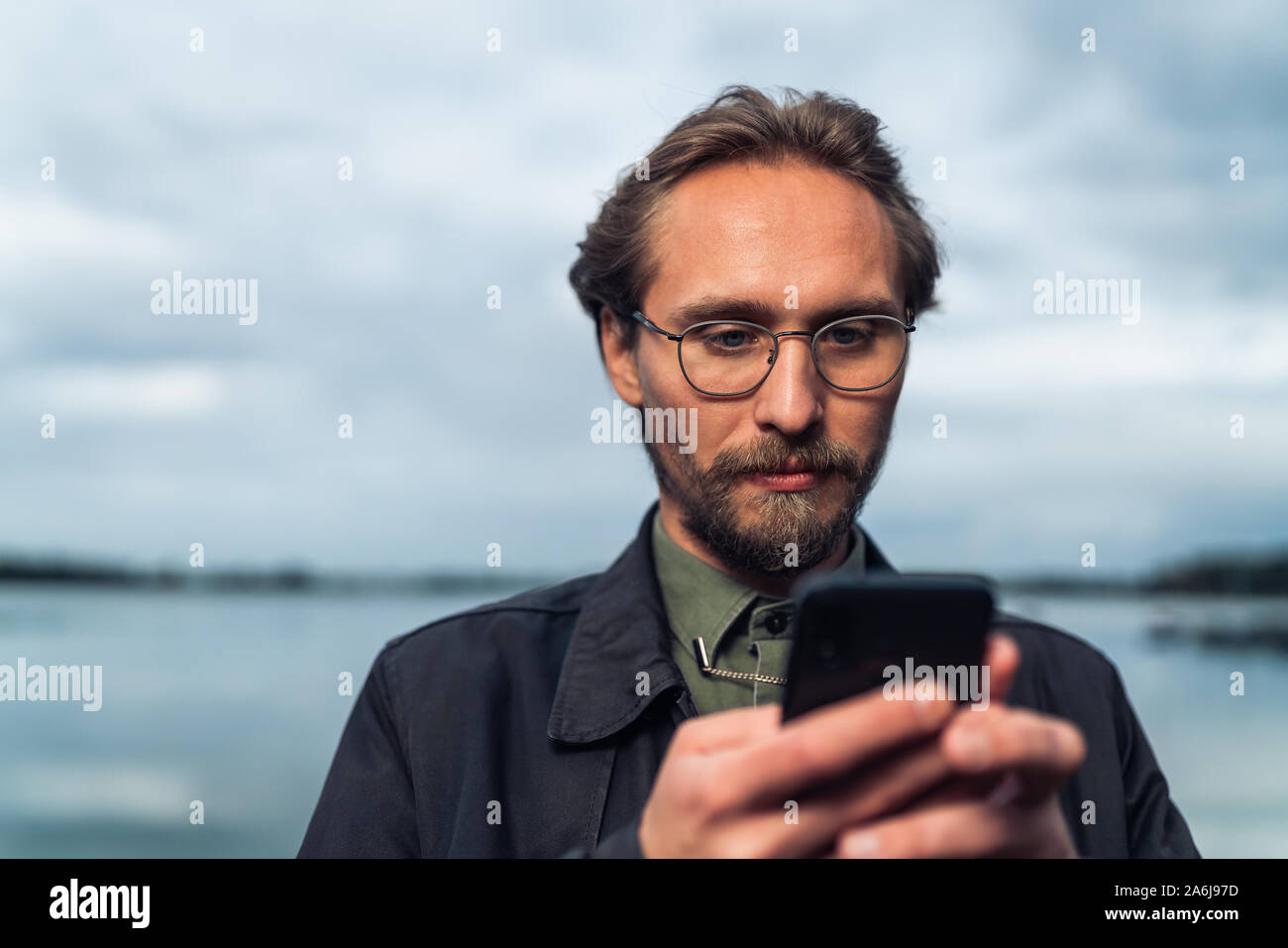 Jungen gutaussehenden Mann auf sein Handy zu schielen. See und Wolken im Hintergrund sichtbar. Stockfoto