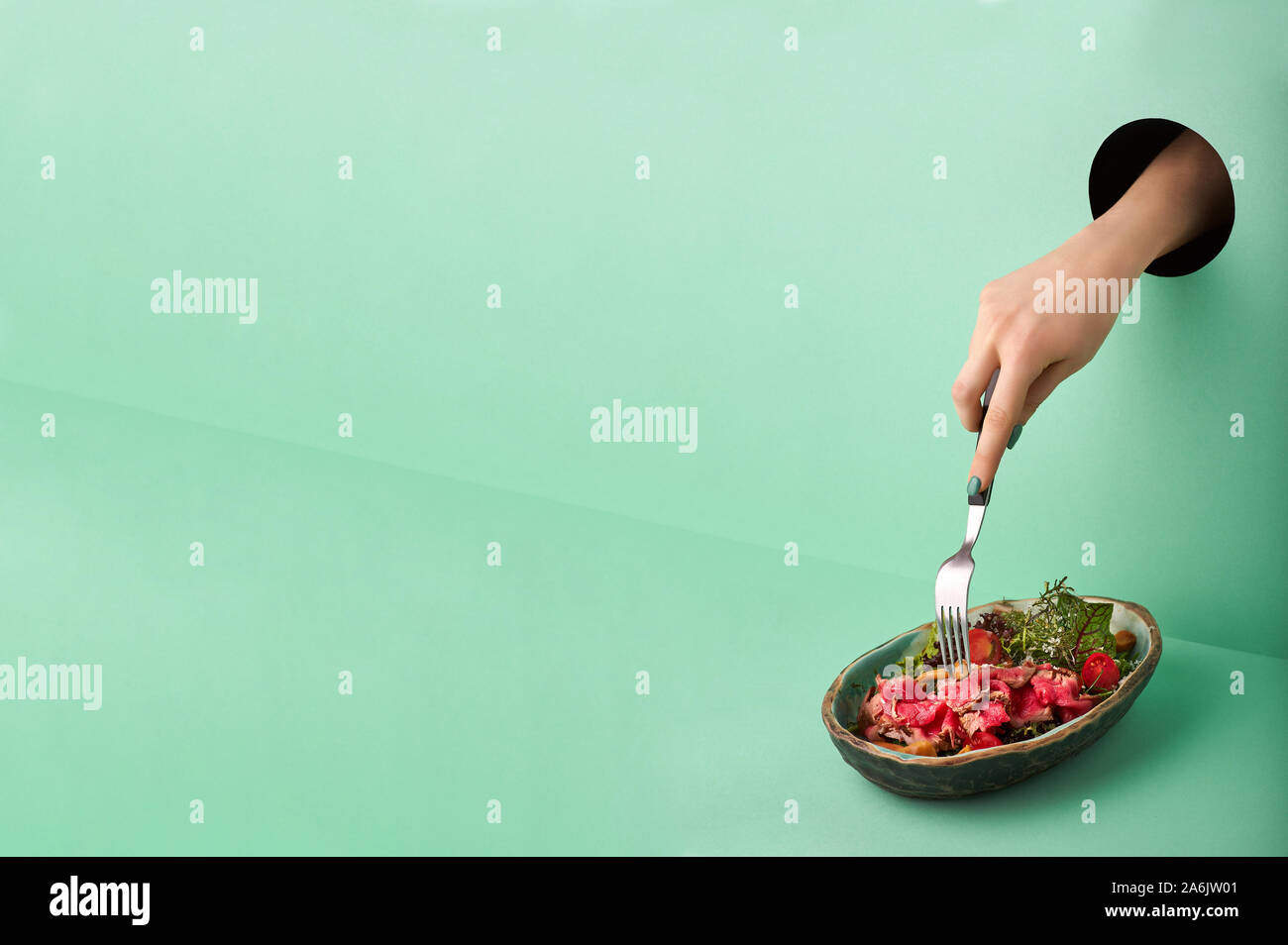 Weibliche Hand reicht für Roastbeef Salat durch das Loch in der grünen Wand. Essen Konzept in der neo-mint Farbe. Hand nimmt Nahrung durch das Loch. Rot Rindfleisch Salat Stockfoto