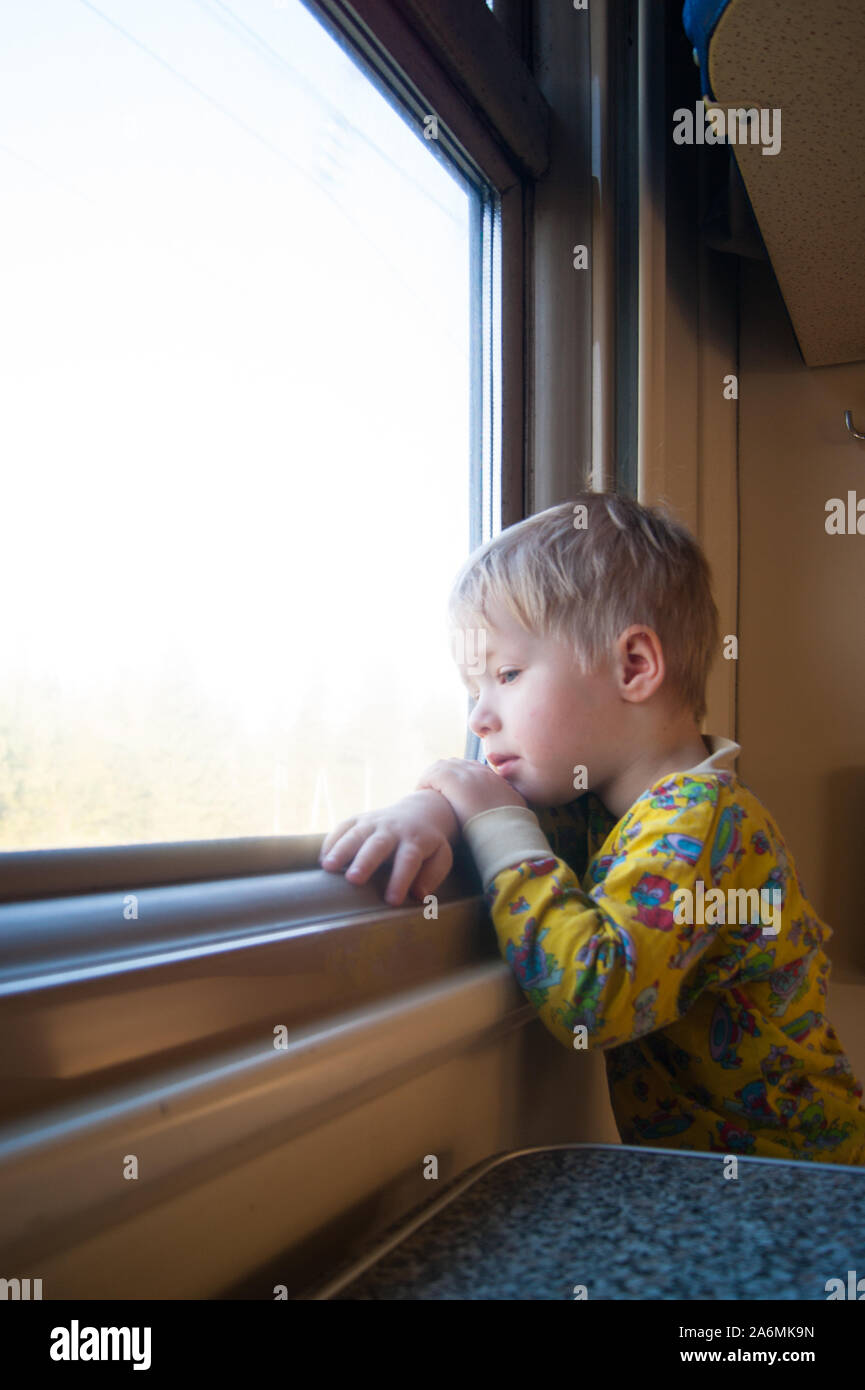 Eine Helle, nette 3-jähriger Junge dem Zug schaut aus dem Fenster, hinter dem sich eine langweilige Landschaft fegt durch. Vertikale Foto. Das Kind ist über etwas. Kind, Kleinkind, niedlich, süß, Neugier, Kognition, Interesse, Reisen, Eisenbahn, Verkehr Stockfoto