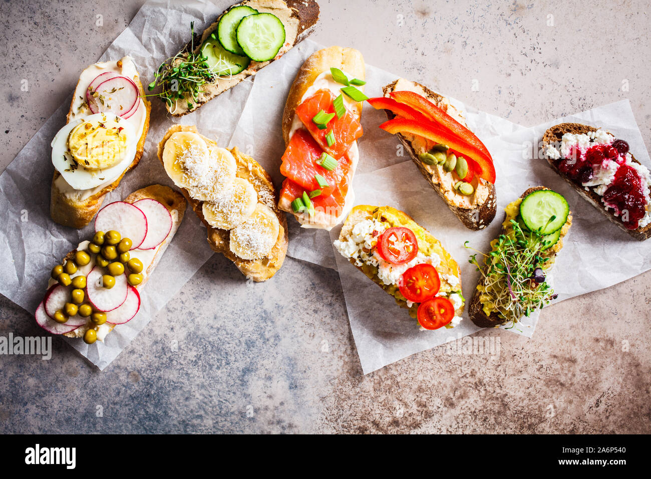 Toast mit verschiedenen Belägen auf grau-braunen Hintergrund geöffnet. Flach von Crostini mit Banane und Erdnussbutter, Pastete, Avocado, Lachs, Ei, Käse Stockfoto