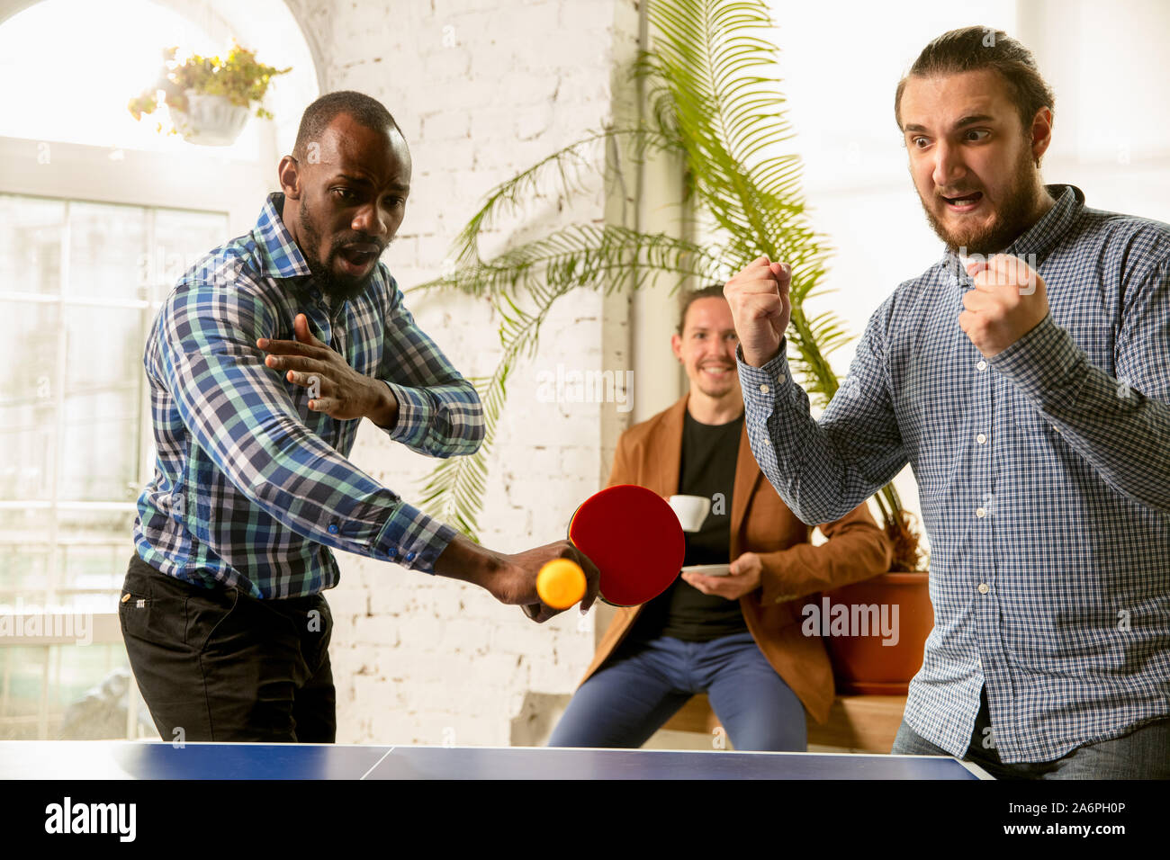 Junge Menschen spielen Tischtennis am Arbeitsplatz, Spaß zu haben. Freunde in legere Kleidung spielen Ping pong zusammen an einem sonnigen Tag. Konzept der Freizeitgestaltung, Sport, Freundschaft, Teambuilding, Teamarbeit. Stockfoto