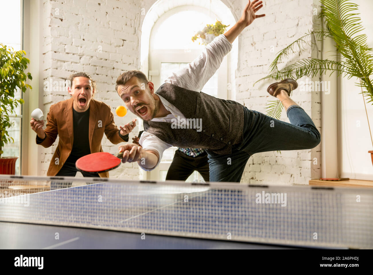 Junge Menschen spielen Tischtennis am Arbeitsplatz, Spaß zu haben. Freunde in legere Kleidung spielen Ping pong zusammen an einem sonnigen Tag. Konzept der Freizeitgestaltung, Sport, Freundschaft, Teambuilding, Teamarbeit. Stockfoto