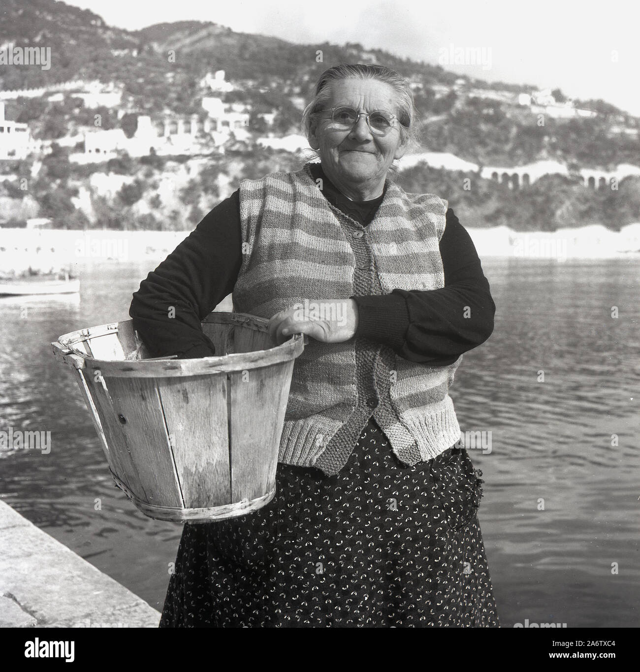 1950er Jahre, historisch, eine ältere französin mit einem Holzeimer an einem Kai an der Mittelmeerküste in Nizza, Frankreich, fotografiert von J. Allan Cash. Stockfoto