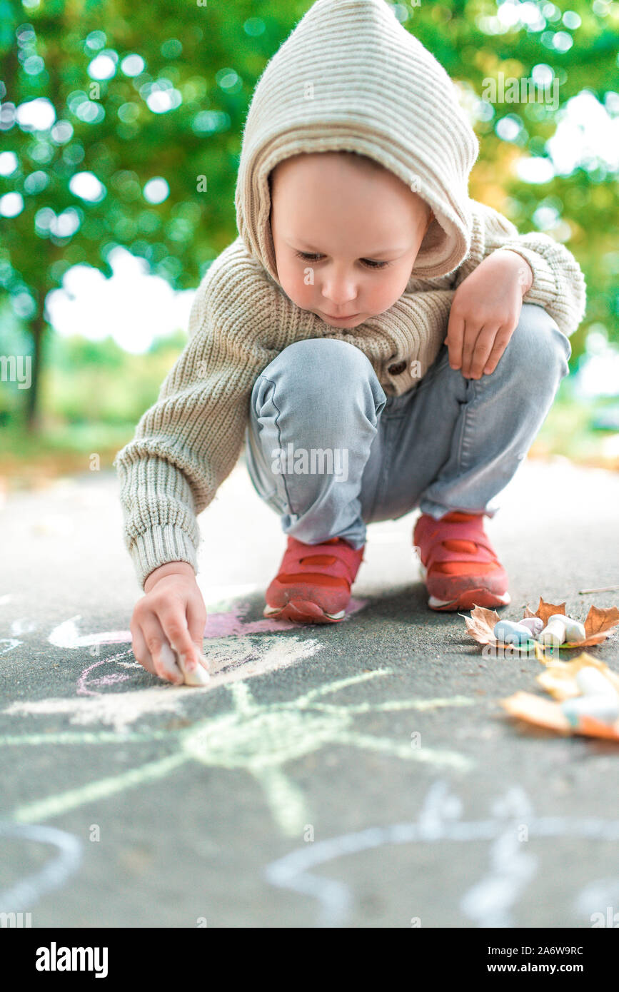 Kleiner Junge Kind von 4-5 Jahre alt, Herbst Tag im Sommer im Stadtpark, zieht mit Buntstiften, spielt er ernst und hat Spaß. Warme Freizeitbekleidung Pullover Stockfoto