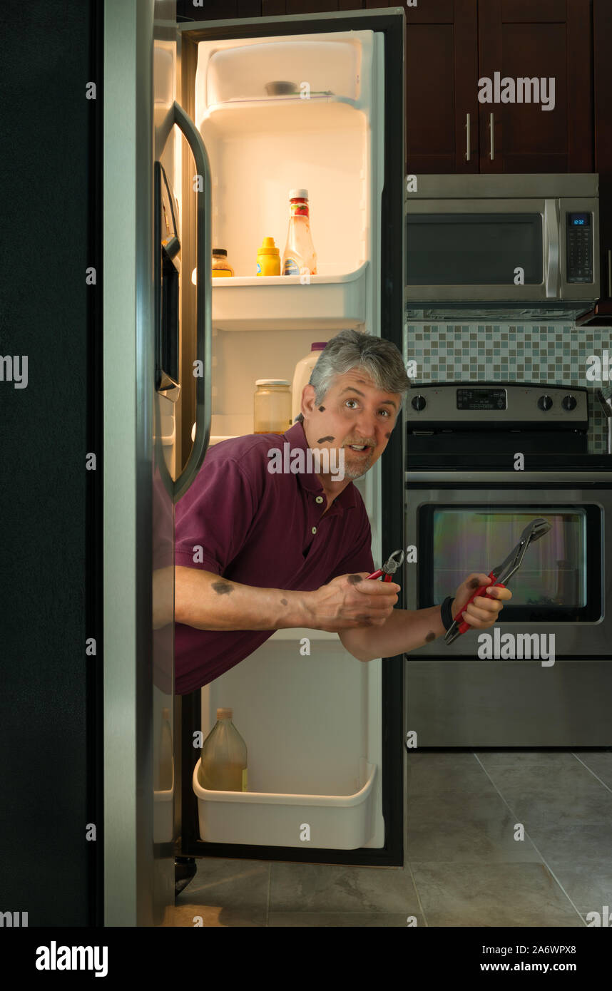 Lustig, humorvoll schmutzig Appliance Reparatur Service Mann oder diy home Eigentümer mit Tools aus einem Kühlschrank hängen in seine Verdatterte versuchen zu korrigieren, showin Stockfoto