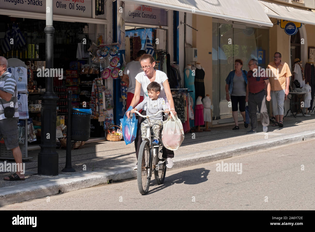 Eine Frau auf einem Fahrrad kehrt nach Hause zurück vom Einkaufen mit ihrem Sohn. Sie ist Ausgleich selbst, ihr Sohn & Einkaufstaschen. In der Griechischen Insel Stadt Kotokolon Stockfoto