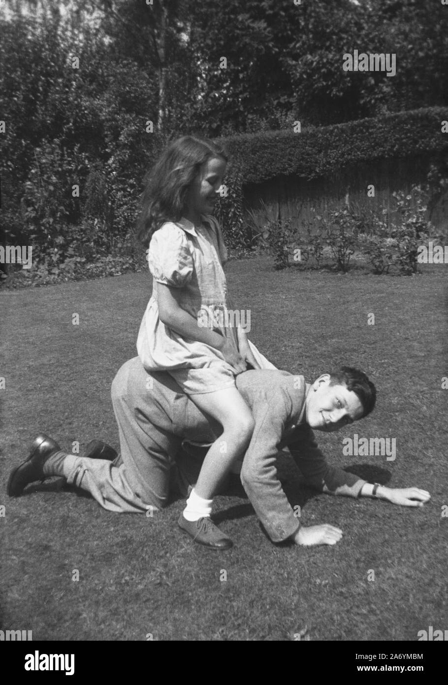 1940er Jahre, historisch, draußen in einem Garten, ein junges Mädchen, das auf dem Rücken ihres älteren Bruders sitzt und so tut, als würde sie ein Pferd reiten, England, Großbritannien. Stockfoto