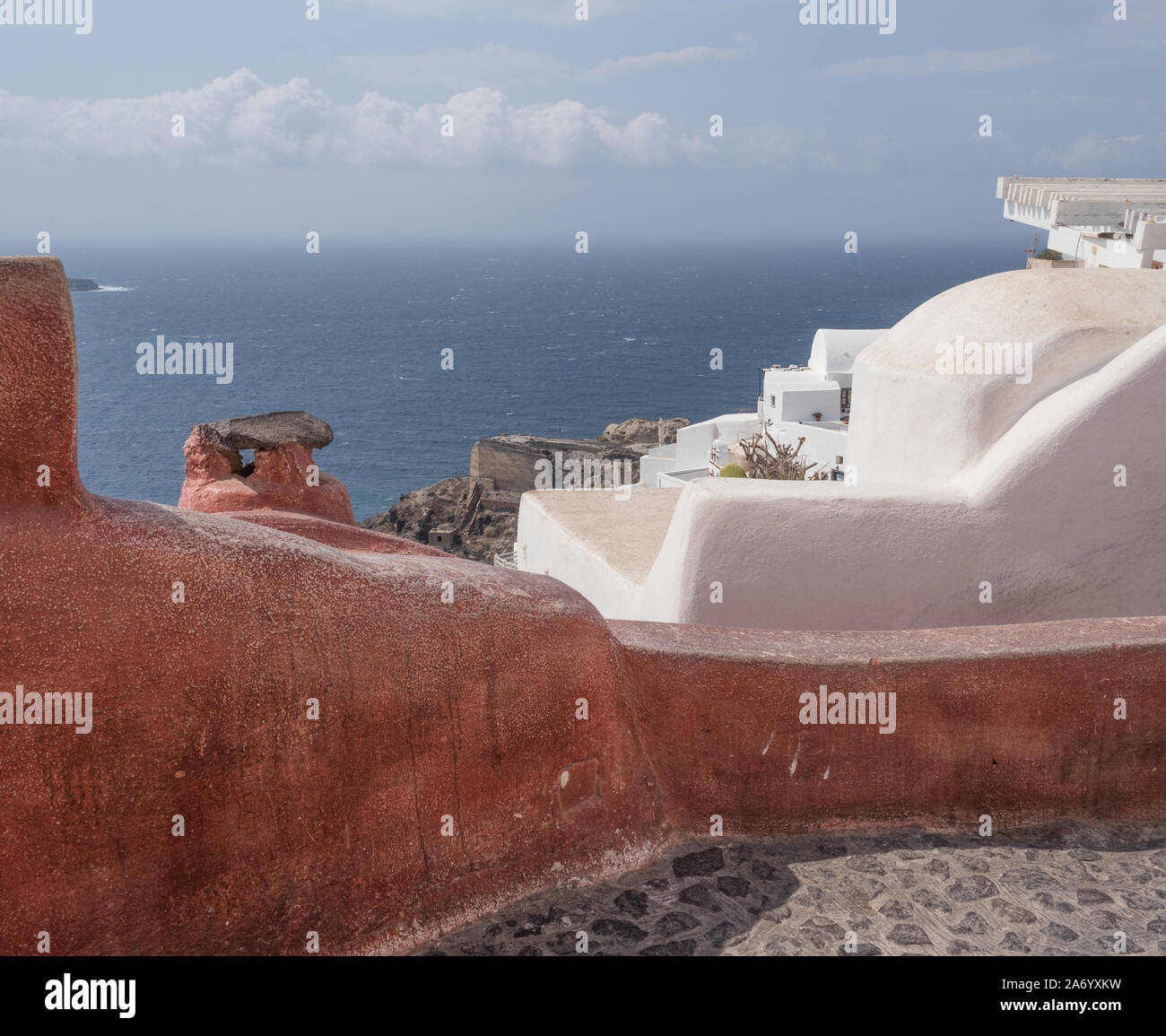 Ein erdiges rote und weiße Wand Wände Kopfsteinpflaster Stein Kopfsteinpflaster Boden mit Bergen im Hintergrund und einem ruhigen Meer unter blauem Himmel mit Wolken Oia Santorini Stockfoto