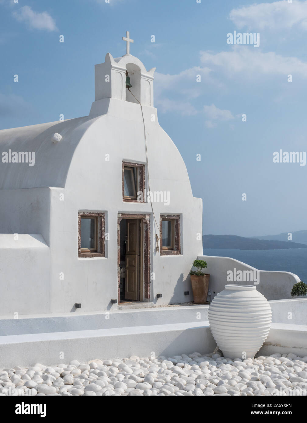 Ein weißer waschen Kapelle Kirche offene Tür kleinen Glockenturm mit Kreuz cruicifix blue sky cloud Topf Kopfsteinpflaster niedrige Mauer Vordergrund Oia Santorini Stockfoto