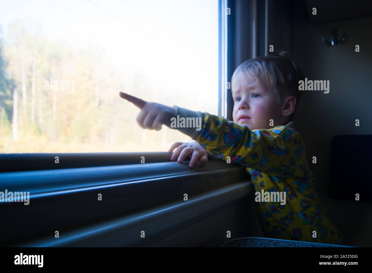 - Der Zug! Eine kleine blonde Junge im Alter von drei Jahren reitet auf einem Zug, isst Bonbons und schaut aus dem Fenster, hinter dem sich ein schöner Herbst Landschaft fliegt. Das Kind ist über etwas. Emotionen, Kind, Kleinkind, niedlich, süß, Neugier, Kognition, Interesse, Reisen, Eisenbahn, Verkehr Stockfoto