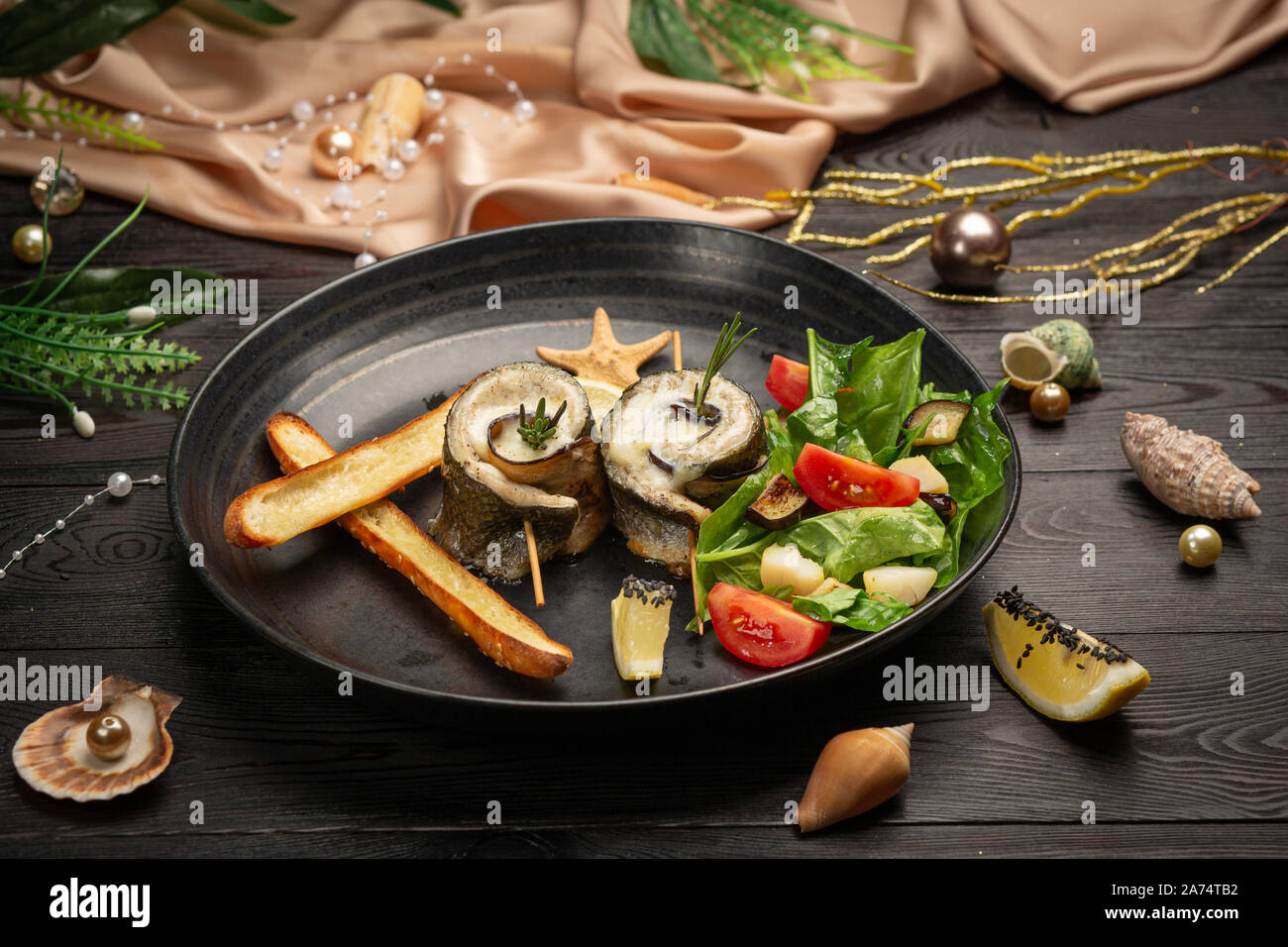 Gebackener Wolfsbarsch mit Auberginen, Mozzarella mit frischem Salat und Grissini Stöcken in einer schwarzen Platte auf einem dunklen Hintergrund Stockfoto