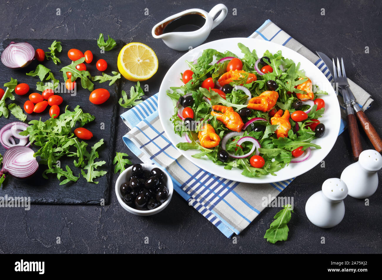 Rucola crab Schellen Salat mit schwarzen Oliven, Tomaten und roten Zwiebeln auf einem weißen Teller serviert, auf einer konkreten Tabelle mit Zutaten, die Aussicht von ein Stockfoto