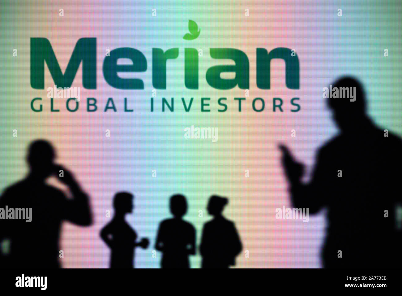 Der Merian Global Investors Logo ist auf einen LED-Bildschirm im Hintergrund, während eine Silhouette Person ein Smartphone verwendet (nur redaktionelle Nutzung) Stockfoto