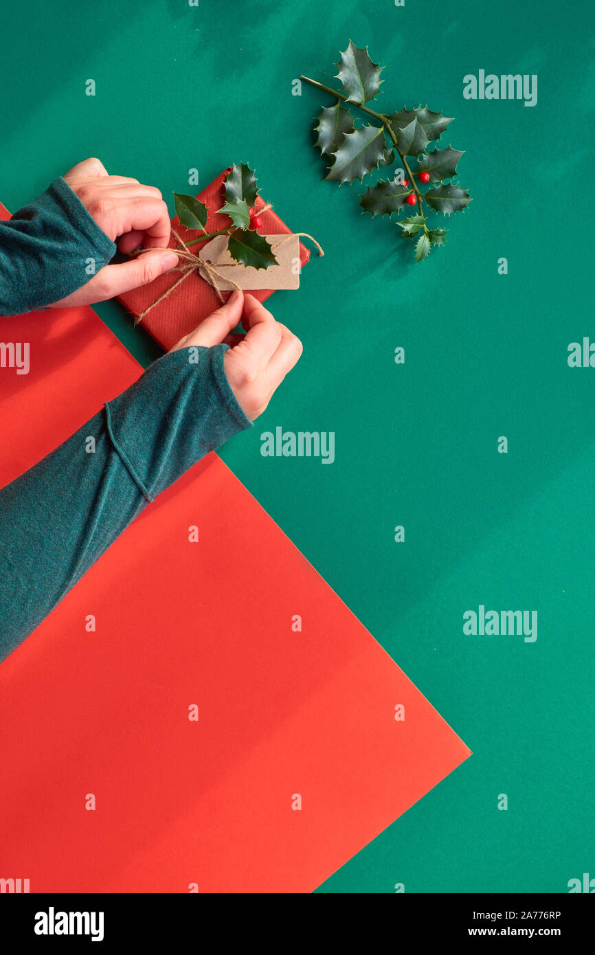 Kreative diagonal Geometrische flach an der grünen und roten Papier mit langen floralen Schatten. Verpackt Geschenkkarton dekoriert mit Holly. Weibliche Hände binden die Stockfoto