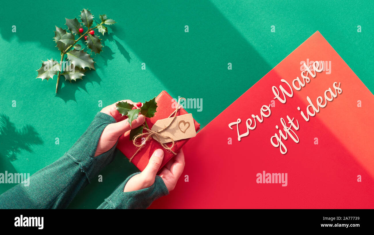 Kreative Panoramablick flach an der grünen und roten Papier mit langen Schatten. Weibliche Hände halten verpackt Geschenkkarton dekoriert mit Holly. Umweltfreundliche Weihnachten, Stockfoto