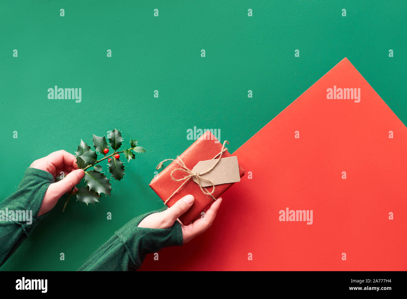 Geometrische flach auf grüne und rote Papier legen Kopieren - Raum. Weibliche Hand gewickelt Geschenkbox und Zweig von Holly mit roten Beeren. Umweltfreundliche Geschenk ide Stockfoto