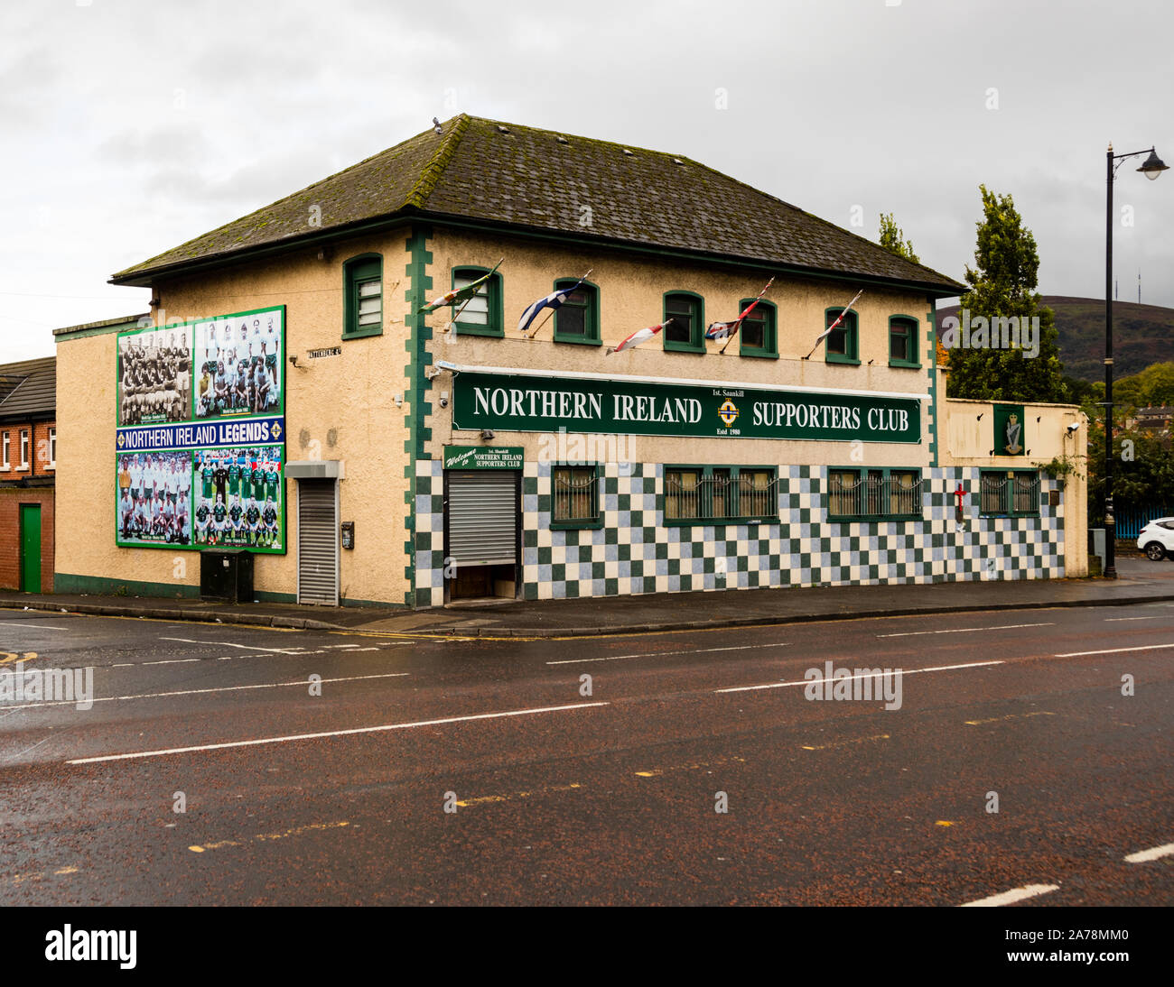 Das Clubhaus des 1st Shankill Northern Ireland Supporters Club an der Shankill Road in Belfast, Nordirland, trägt an seiner Fassade das Wandgemälde „Northern Ireland Legends“ mit Teamfotos der nordirischen Nationalfußballmannschaft; ein grün-weißes Schachbrettmuster schmückt das Erdgeschoss, während Clubflaggen an den Fenstern des Obergeschosses hängen. Stockfoto