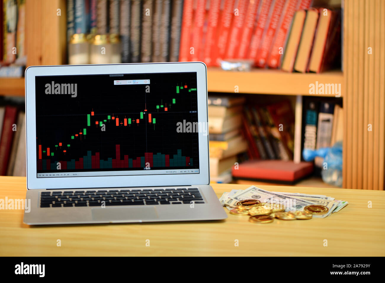 Auf dem Tisch golden Bitcoins und Laptop mit Börse Grafik auf dem Bildschirm, verschwommenes Regal mit Büchern in der background-image Stockfoto