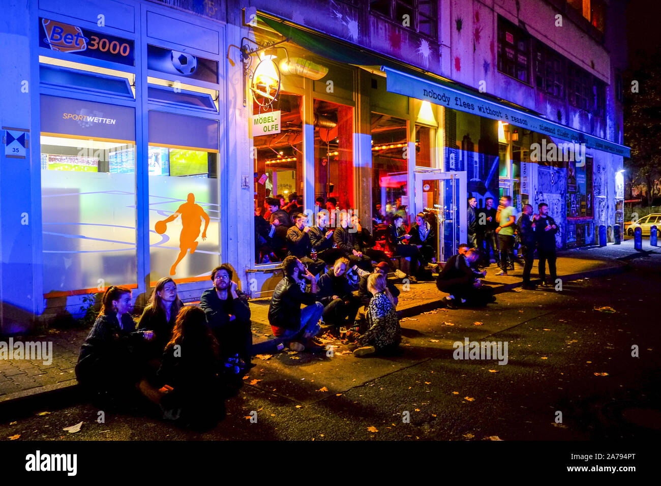 Berlin, Deutschland. 12 Okt, 2019. Junge Menschen sitzen auf der Straße vor  der Bar 'Möbel Olfe" am Kottbusser Tor im Bezirk Kreuzberg, während die  Fassade blau durch das blaue Licht von einem