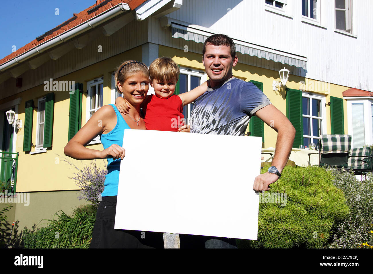 Junge Familie mit Art vor neugekauftem Haus, hält weisses Schild, frei für beliebigen Text, 30,35, 5, Jahre, HERR: Ja Stockfoto