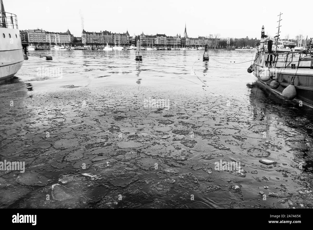 Schwarz-weiß-Bild des Stockholmer River Harbour in der Innenstadt mit zwei Booten an den Seiten, Eiswasser im Vordergrund und die Altstadt in der Backgr Stockfoto