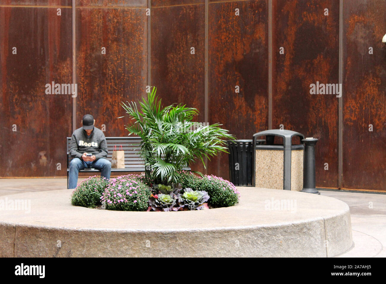 Ein Mann sitzt auf einer Bank hinter einer Pflanzmaschine und vor einem hohen rostige Wand auf der Außenseite eines Condo-gebäude Stockfoto