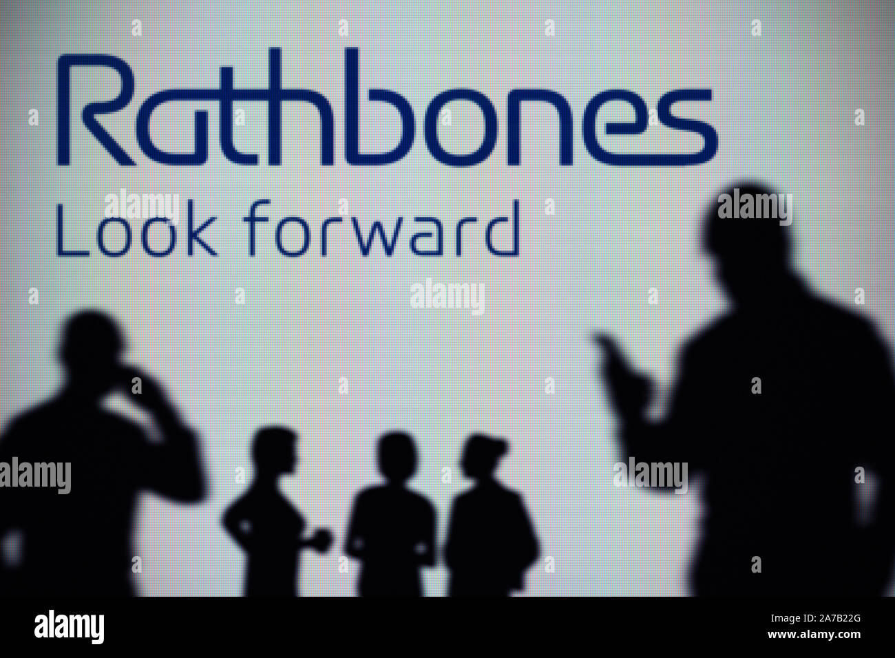 Das Rathbone Brothers Logo ist auf einen LED-Bildschirm im Hintergrund, während eine Silhouette Person ein Smartphone verwendet (nur redaktionelle Nutzung) Stockfoto