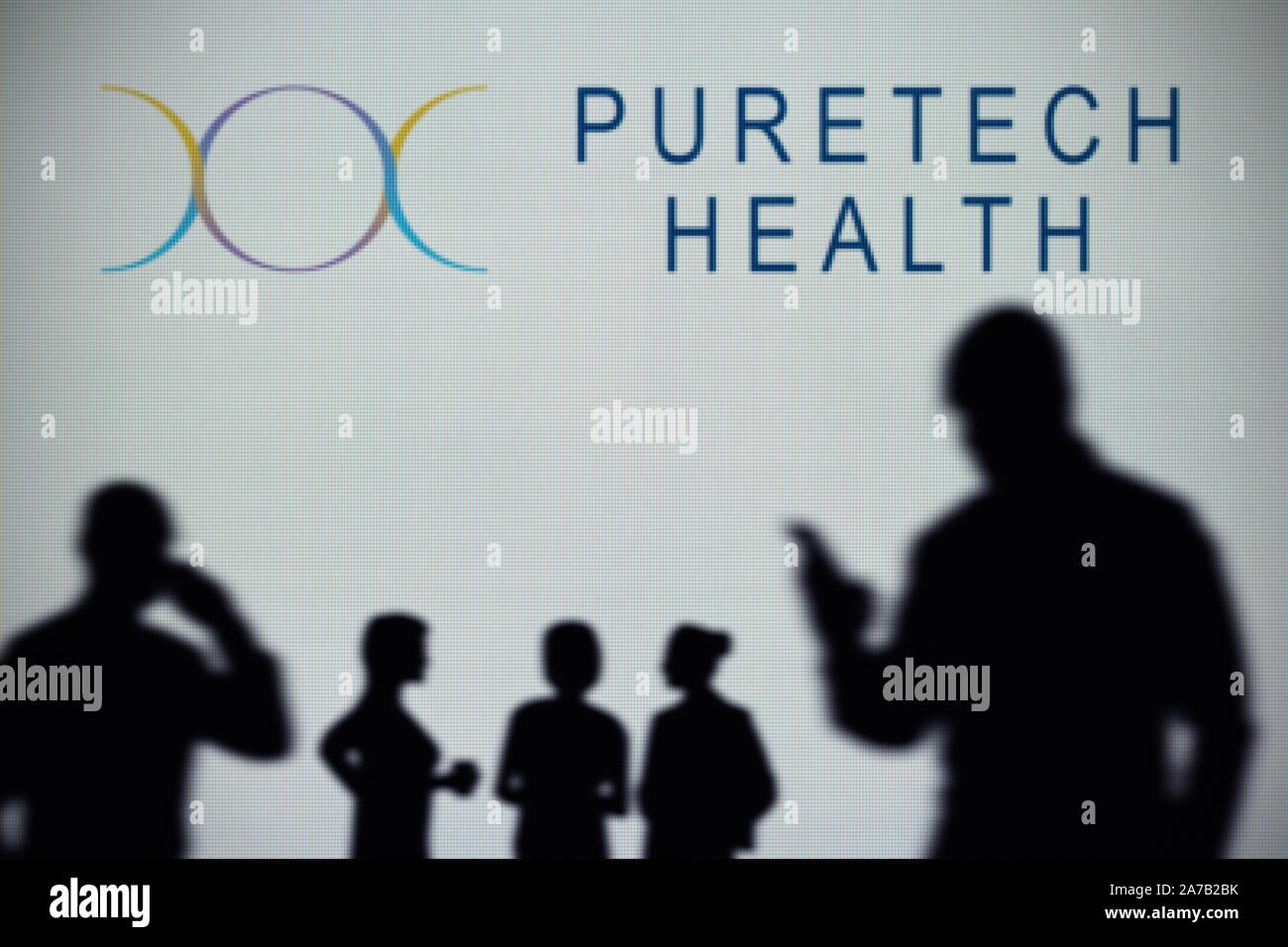 Die Puretech Gesundheit Logo ist auf einen LED-Bildschirm im Hintergrund, während eine Silhouette Person ein Smartphone verwendet (nur redaktionelle Nutzung) Stockfoto