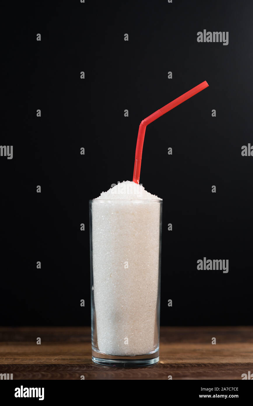 Ein Glas voll mit weißem Zucker mit Stroh auf schwarzem Hintergrund. Konzept der ungesunden Ernährung und Diabetis. Bild vertikal Stockfoto