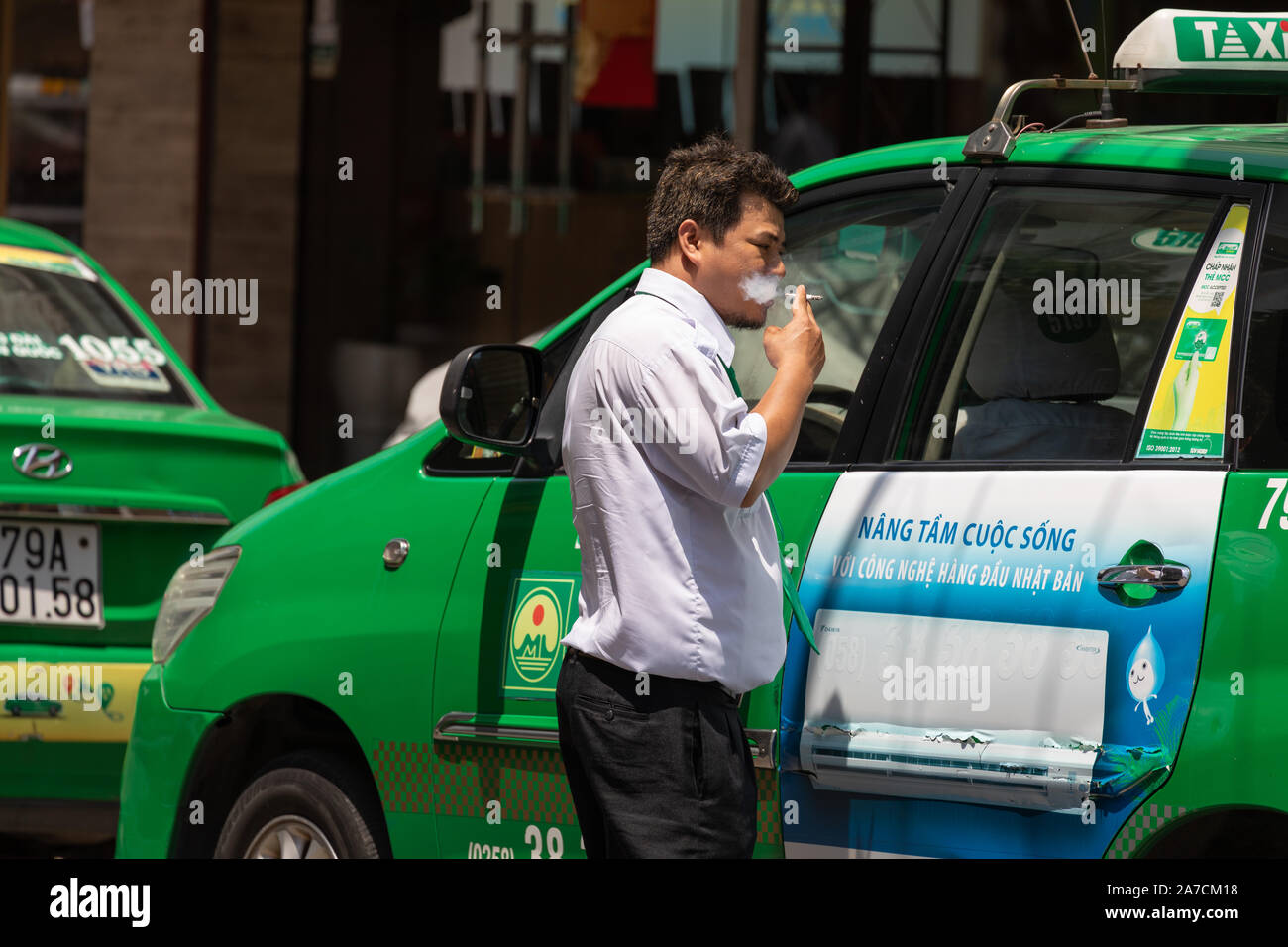 Vietnam, Nha Trang City, 25. März 2019 Eine vietnamesische Taxifahrer raucht nervös, während außerhalb seines grünen Taxis warten Stockfoto
