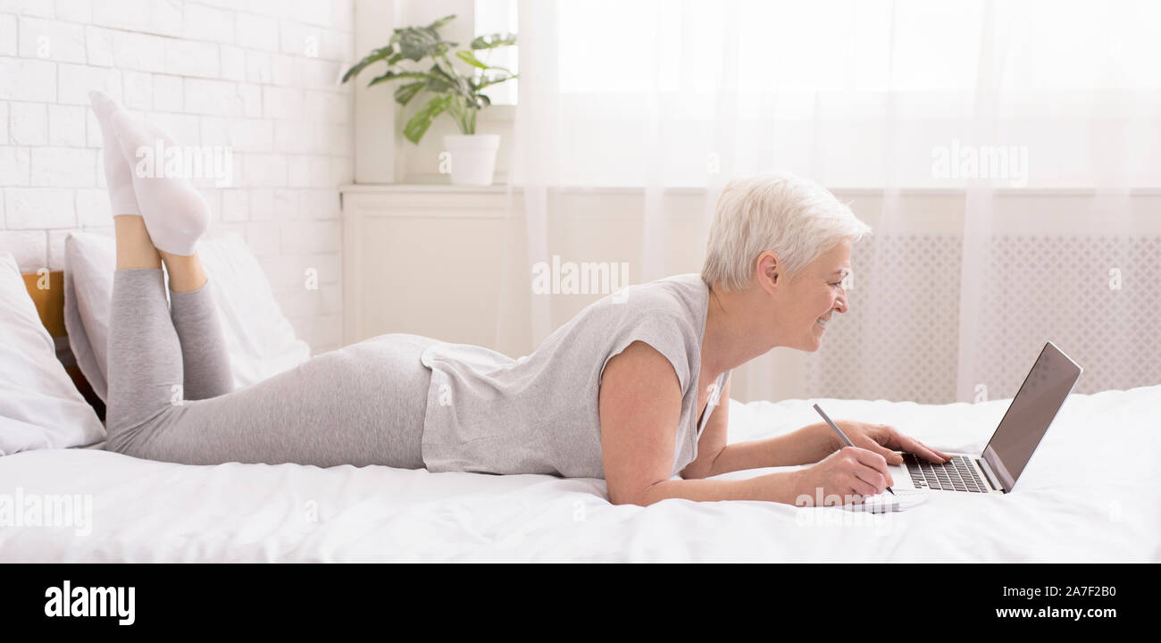 Freundliche ältere Dame plaudern auf Laptop online, liegend auf Bett Stockfoto