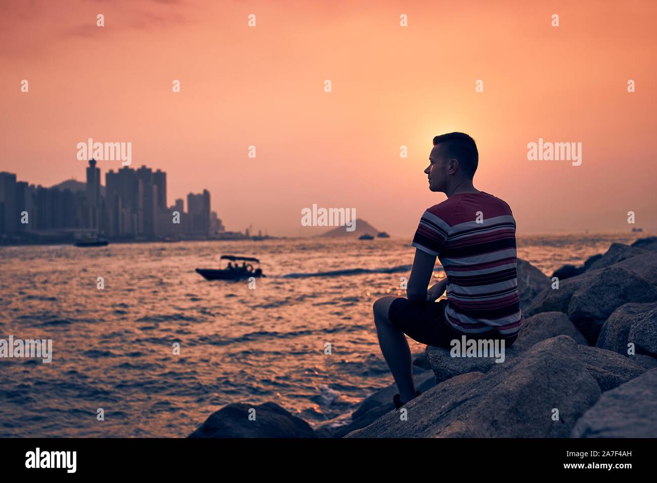Junge Mann sitzt auf Steine gegen Hong Kong Stadtbild mit seinen Wolkenkratzern im Golden Sunset. Stockfoto