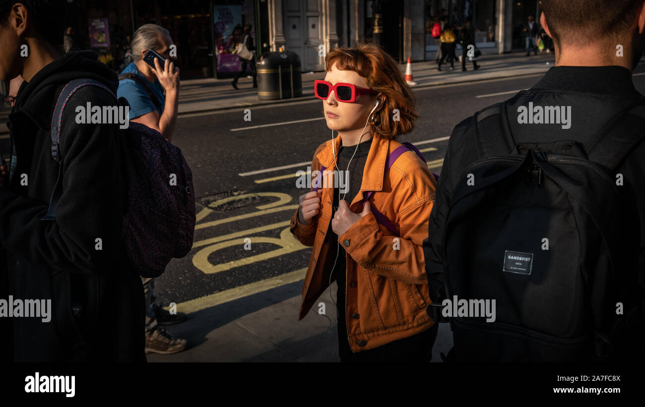 Eine junge Frau macht, an der Londoner Oxford Street mit der tief stehenden Sonne Kommissionierung ihre rote Sonnenbrille durch die Menge Stockfoto