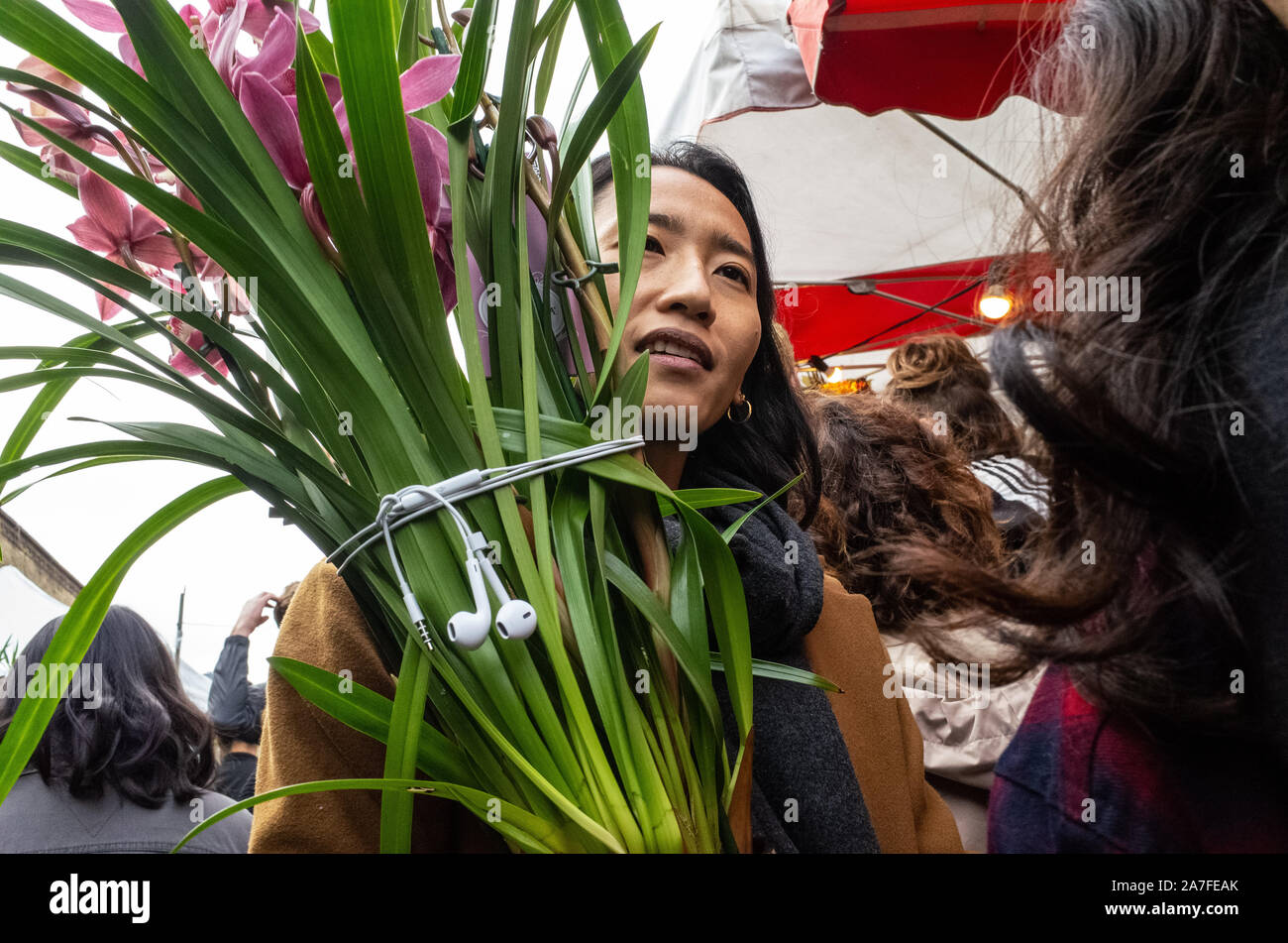 Asiatische Dame können Sie über Ihre Kopfhörer eine Pflanze zu unterstützen, wie sie trägt es durch Columbia Road Blumenmarkt, East London, Großbritannien Stockfoto