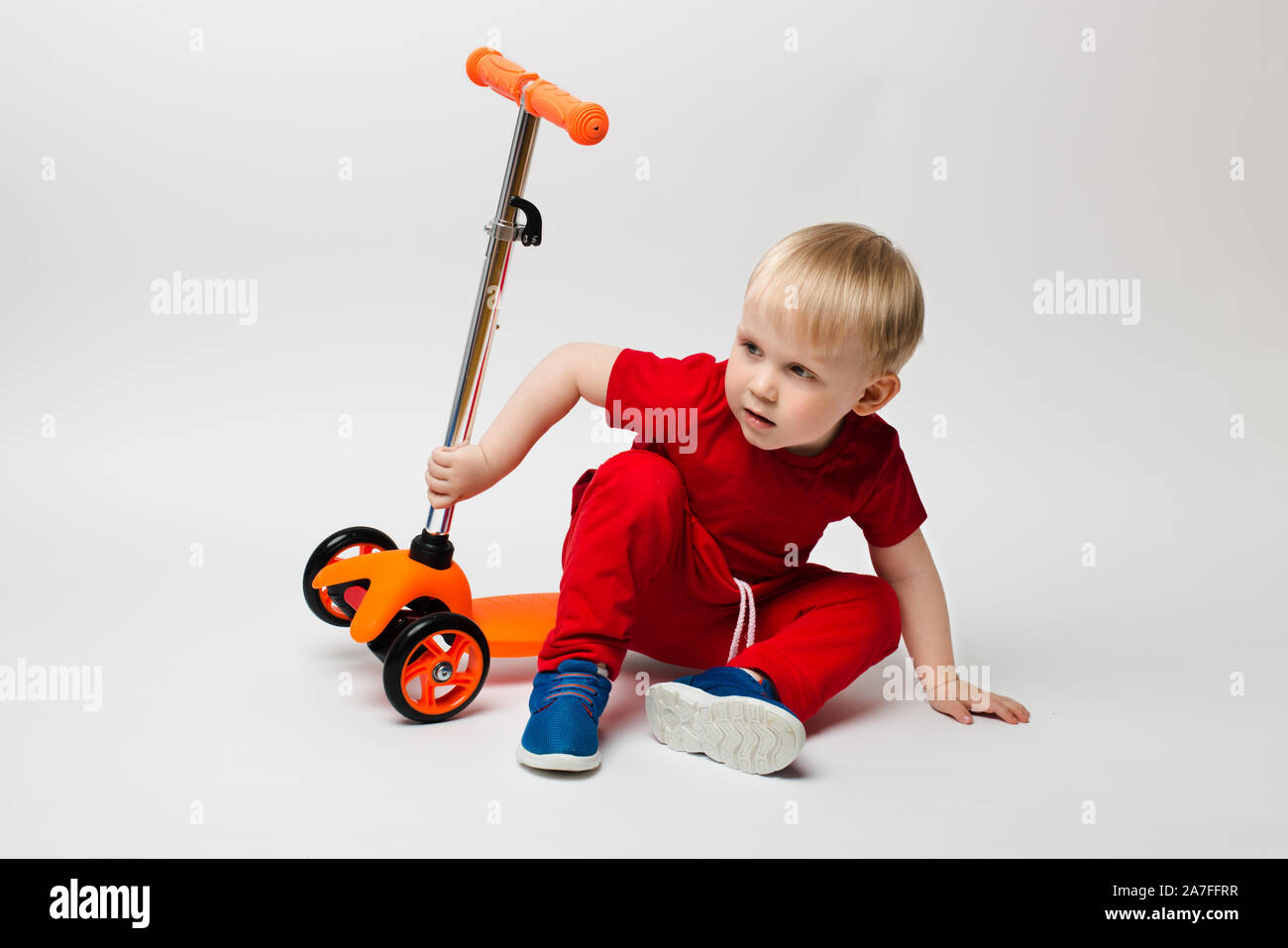 Niedliches Kleinkind Kind in ein rotes T-Shirt fiel von einem Roller und sitzt, Aufnahmen im Studio auf einem weißen Hintergrund. Konzept für Artikel über Kindheit ein Stockfoto