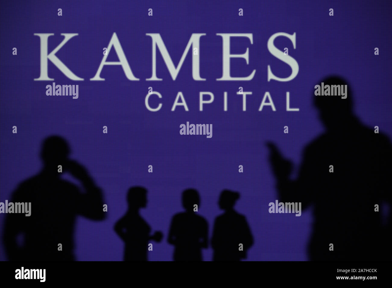Die kames Capital plc-Logo ist auf einen LED-Bildschirm im Hintergrund, während eine Silhouette Person ein Smartphone verwendet (nur redaktionelle Nutzung) Stockfoto