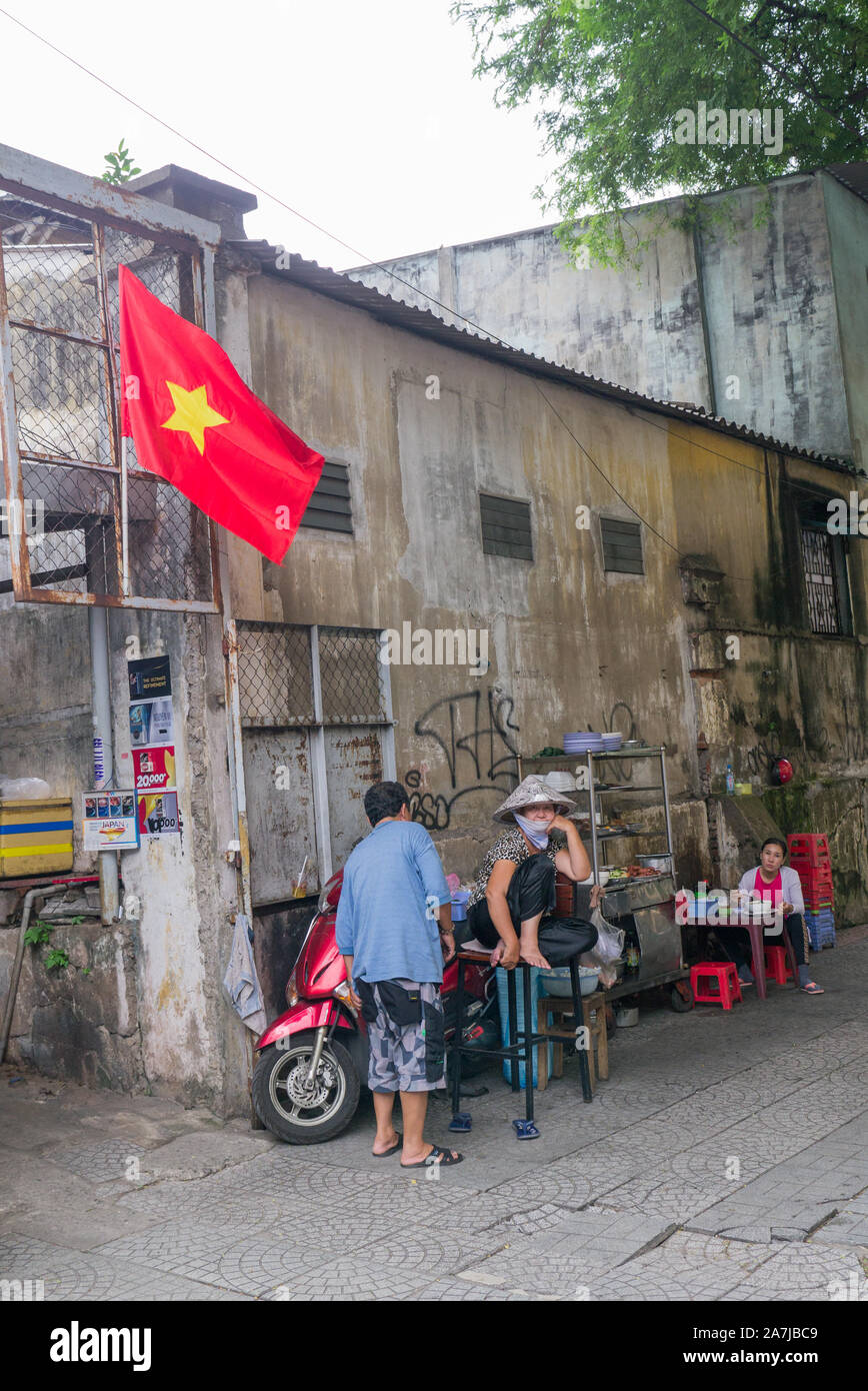 Saigon Seitenszene mit leuchtend roter Vietnam-Flagge und lokalen Straßenverkäufern, die Roller parken, die Frau, die lächelnd auf dem Stuhl sitzt und die Kamera betrachtet. Stockfoto