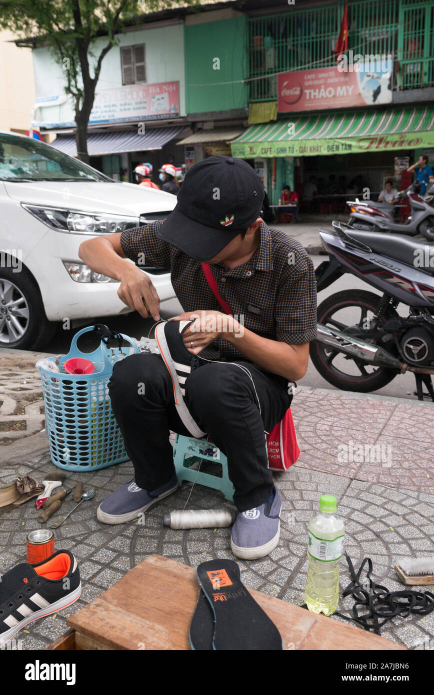 Ein junger Vietnamese, der eine schwarze Baseballkappe trägt, sitzt auf Holzhocker auf dem Gehweg und repariert ein Paar schwarzer Sneaker. Stockfoto